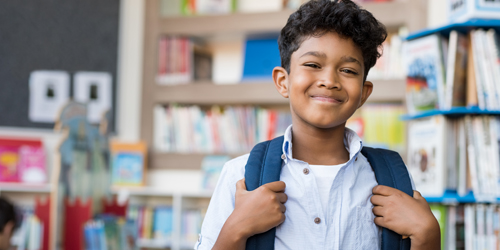 Young elementary schoolboy carrying backpack and standing in library at school. 